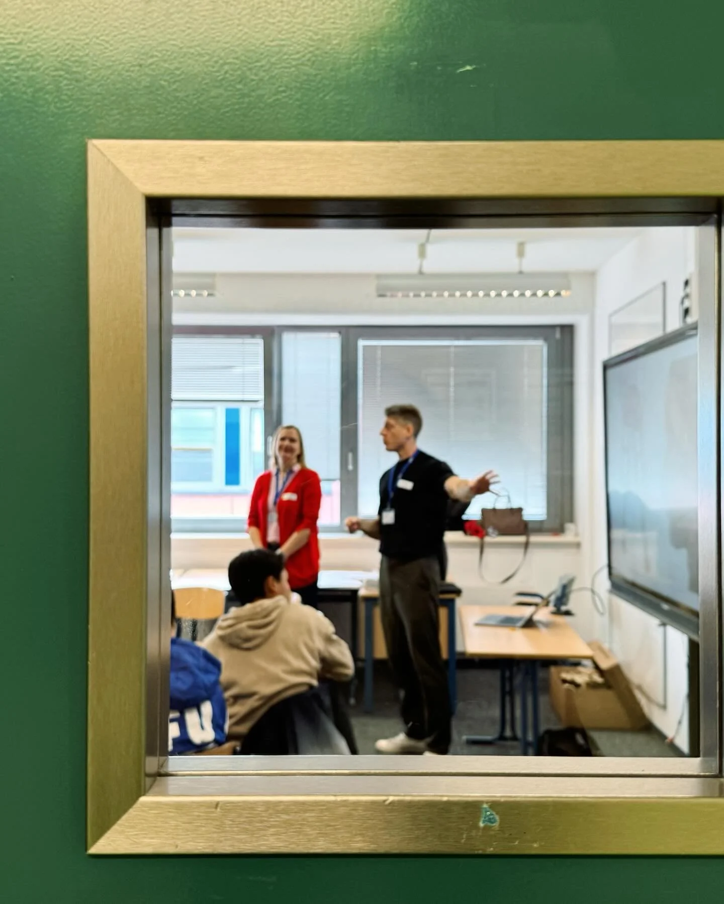 View through a glass window into a classroom where two facilitators stand at the front, engaging with seated participants.